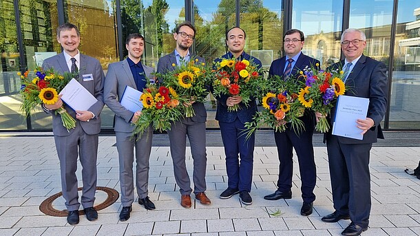 Sechs Männer in Anzügen stehen vor einem verspiegelten Gebäude. Jeder hält einen Blumenstrauß und eine Urkunde in den Händen.